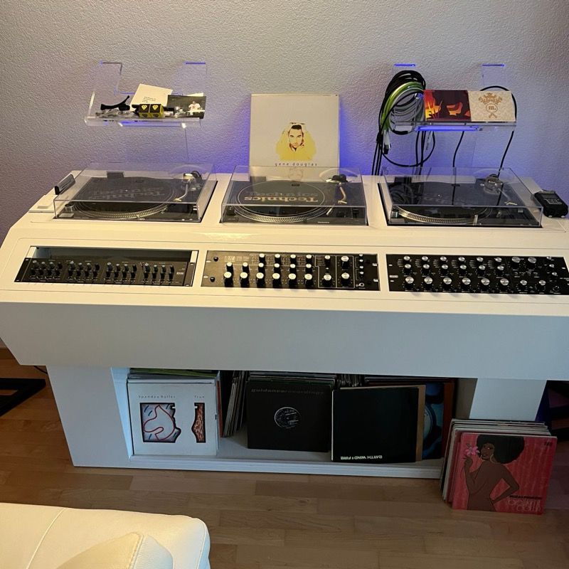 DJ setup with three turntables and mixers in a white console, records stored below