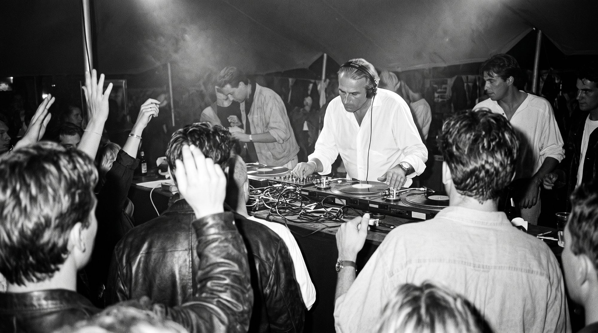 Black-and-white photo of a DJ at the turntables, surrounded by a dancing crowd